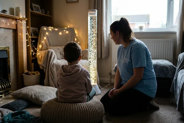 A child seen from behind on a beanbag watching a bubble tube lamp in a softly lit sensory corner, a support worker kneeling alongside