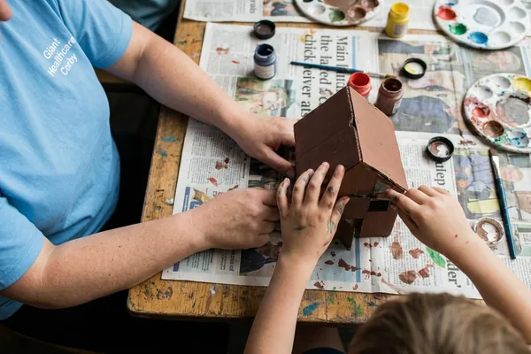 A child's hands and a support worker's hands painting a cardboard model together at a table during an outreach session