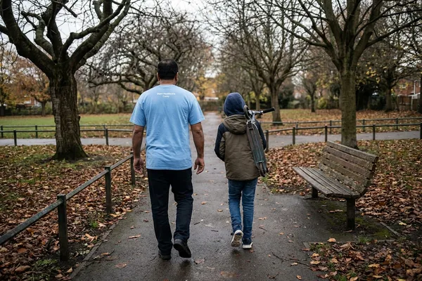A support worker walking through a park with a child carrying a scooter, both seen from behind, on an outreach support session